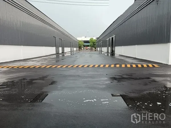Warehouse for rent in Khlong Toei, Bangkok - Wide driveway between two modern warehouse blocks with a yellow speed bump and wet asphalt after rain.