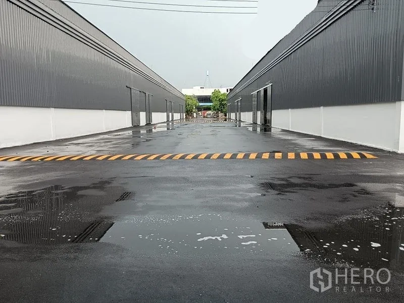 Warehouse for rent in Khlong Toei, Bangkok - Wide driveway between two modern warehouse blocks with a yellow speed bump and wet asphalt after rain.