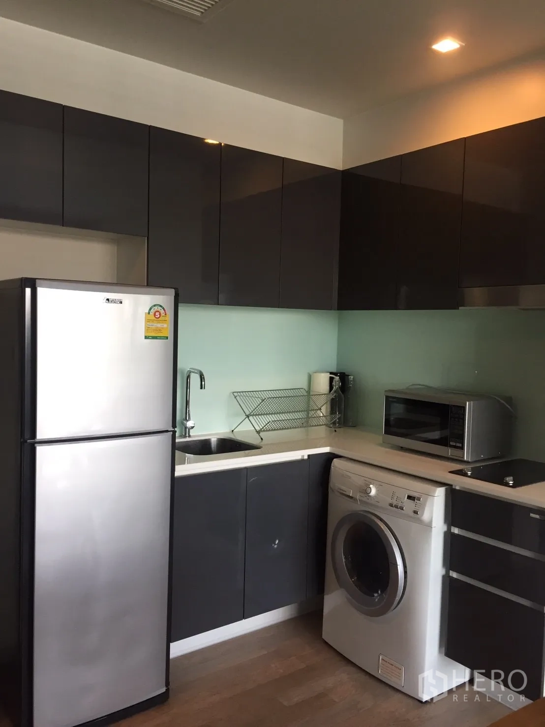 Condominium for rent or sale in Watthana, Bangkok - Close-up of L-shaped kitchen with fridge, washing machine, microwave and glass backsplash.