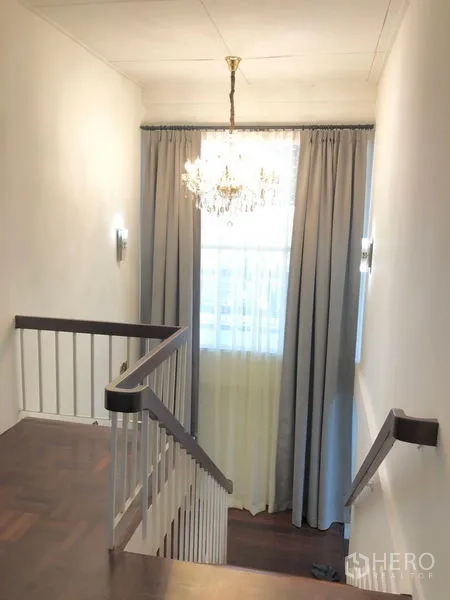 Detached House for rent in Chatuchak, Bangkok - Stair landing with white railing, chandelier, tall window, and long curtains.