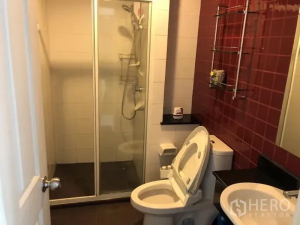 Condominium for rent in Khlong Toei, Bangkok - Bathroom with glass-enclosed shower, toilet, and red accent tile wall.