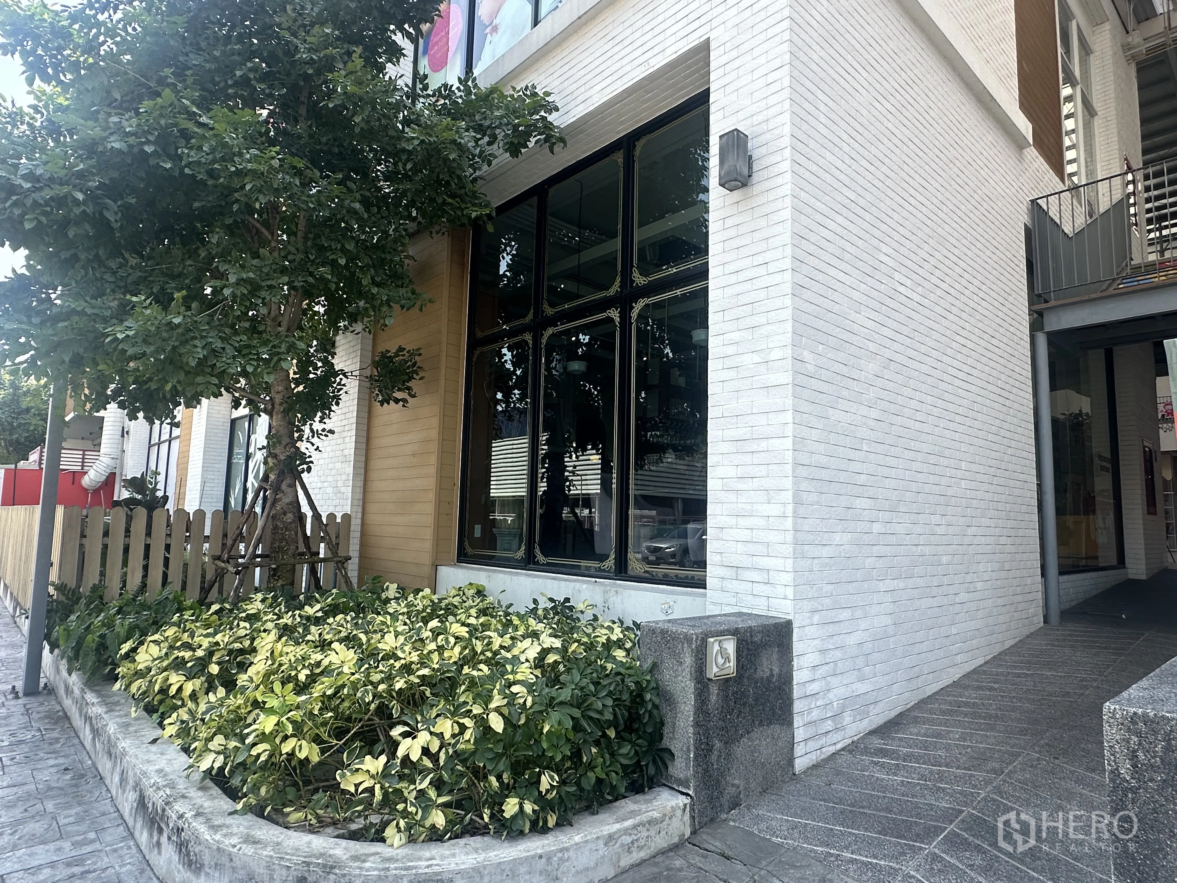 零售空间 for sale in Khlong Toei, Bangkok - 白色砖墙建筑转角，挑高落地窗，配有绿植花坛、树木与侧向通道。