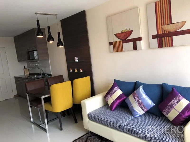 Condominium for rent in Khlong Toei, Bangkok - Open-plan living and dining area with a sofa, colorful cushions, pendant lights, and compact kitchen cabinetry.