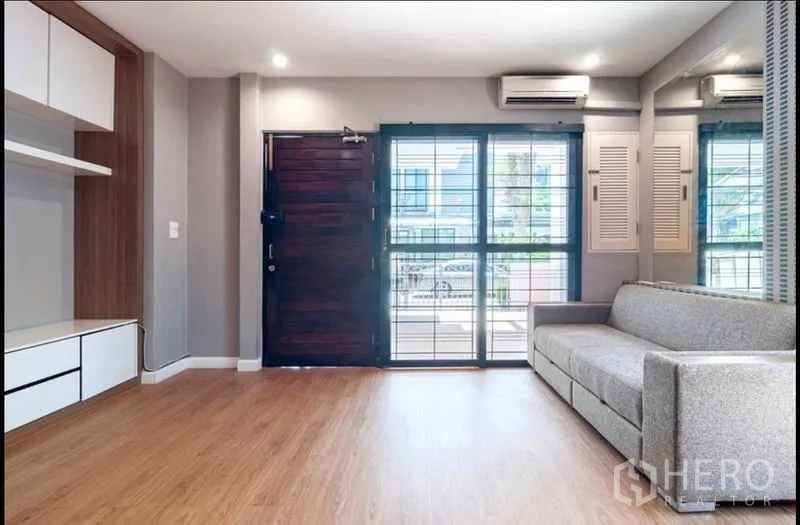 Townhouse for sale in Khlong Toei, Bangkok - Bright living room with wood flooring, large windows, air-conditioning and a grey sofa by the entrance.