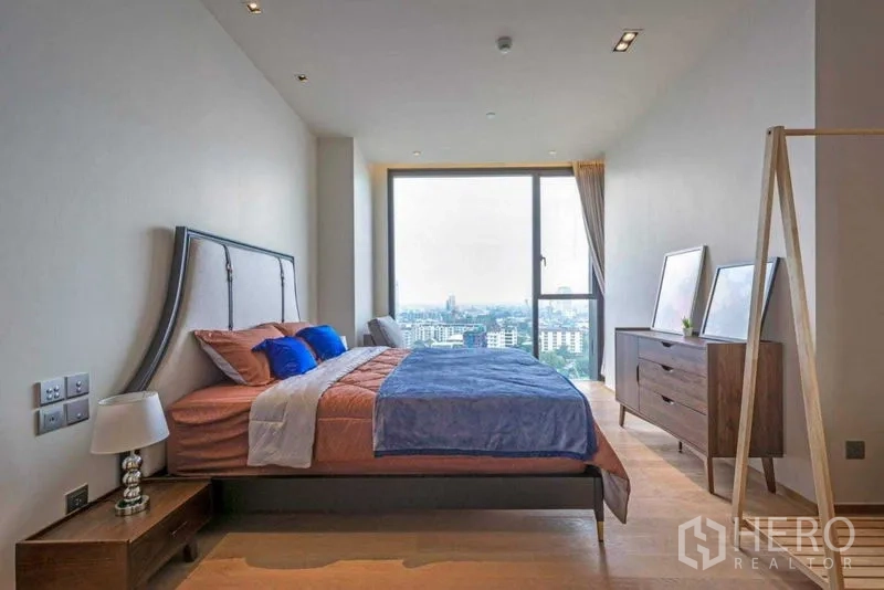 Condominium for rent in Khlong Toei, Bangkok - Bright 1-bedroom condo bedroom with floor-to-ceiling window, city skyline view, wood floors and modern furnishings.