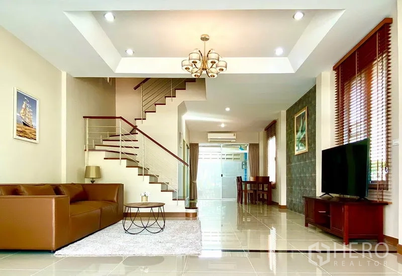 Townhouse for rent in Prawet, Bangkok - Bright open-plan living room with chandelier, staircase, TV console and large windows.