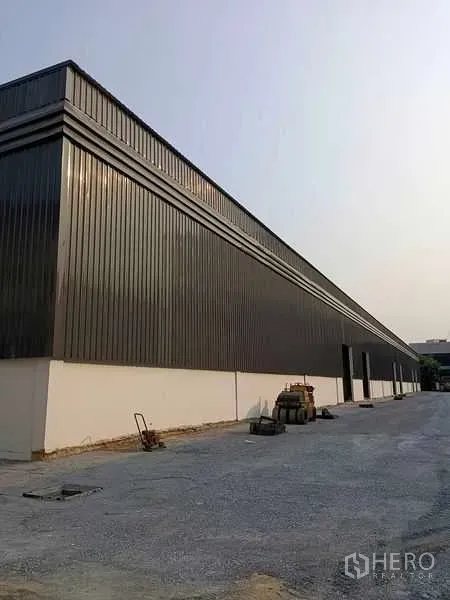 Warehouse for rent in Khlong Toei, Bangkok - Side elevation of a long gray metal-clad warehouse with open yard and equipment along the facade.