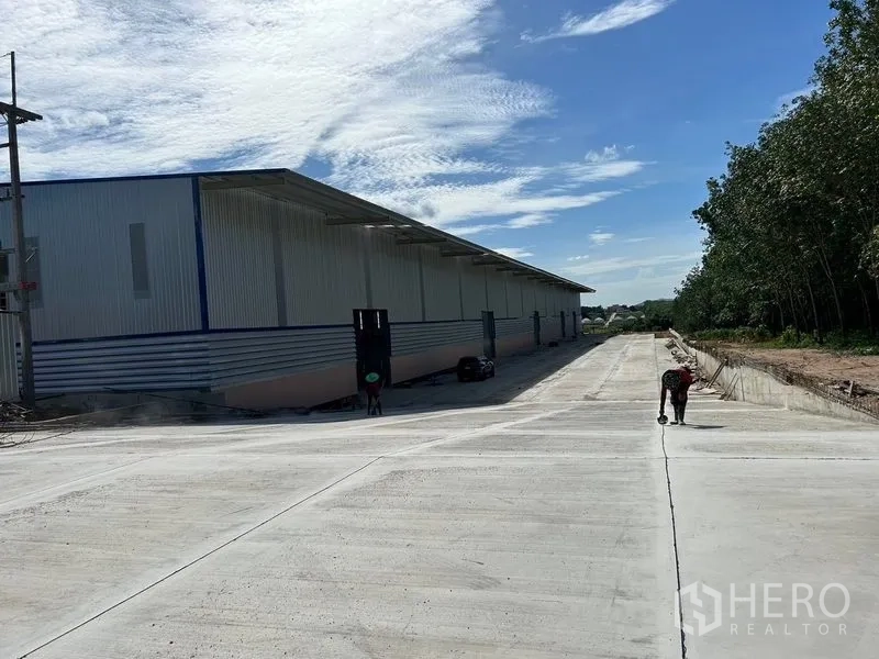 货栈 出租在Nikhom Phatthana, Rayong - 仓库长立面，设有有盖装卸区与宽阔混凝土车道，方便卡车通行。