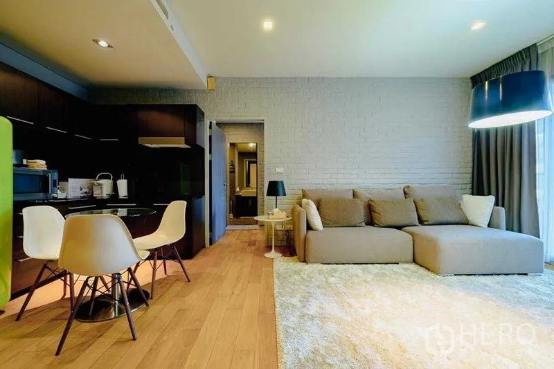 Condominium for rent in Watthana, Bangkok - Open-plan living room with large sectional sofa, wood floors, brick accent wall, and kitchenette with dining set.