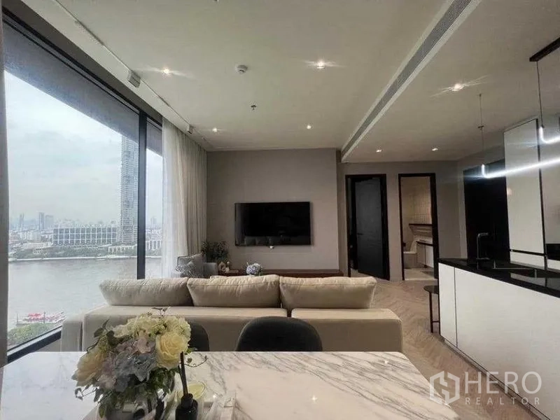 公寓 出租在Khlong San, Bangkok - 开放式客厅，落地窗俯瞰昭披耶河，配沙发、壁挂电视与现代厨房。