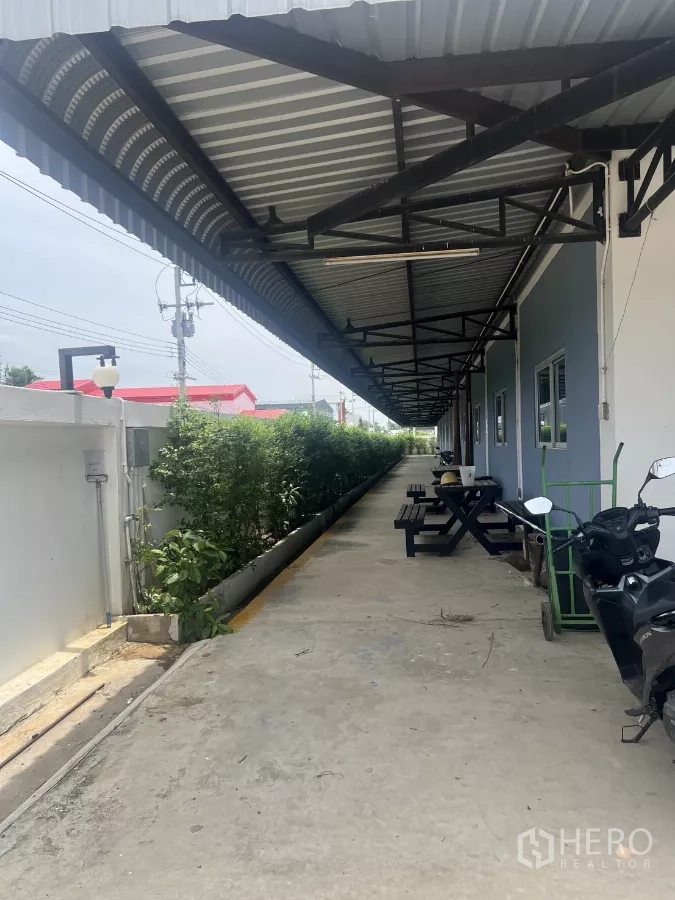 Factory for sale in Lat Lum Kaeo, Pathum Thani - Covered walkway with metal awning, benches, and landscaped greenery beside the building.