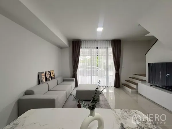 Townhouse for rent in Bang Phli, Samut Prakan - Bright living room with large grey sofa, floor-to-ceiling curtains, TV wall and staircase.