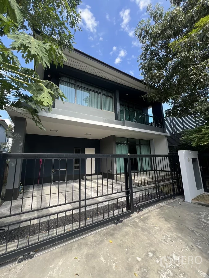 Detached House for rent in Bang Kapi, Bangkok - Modern two-storey detached house exterior with sliding gate, balcony, and driveway shaded by trees.