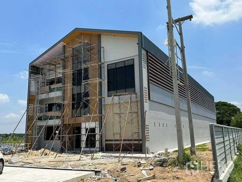 Factory for sale in Khlong Luang, Pathum Thani - Modern factory exterior under construction with scaffolding, gable roof and roller shutter on a fenced site.