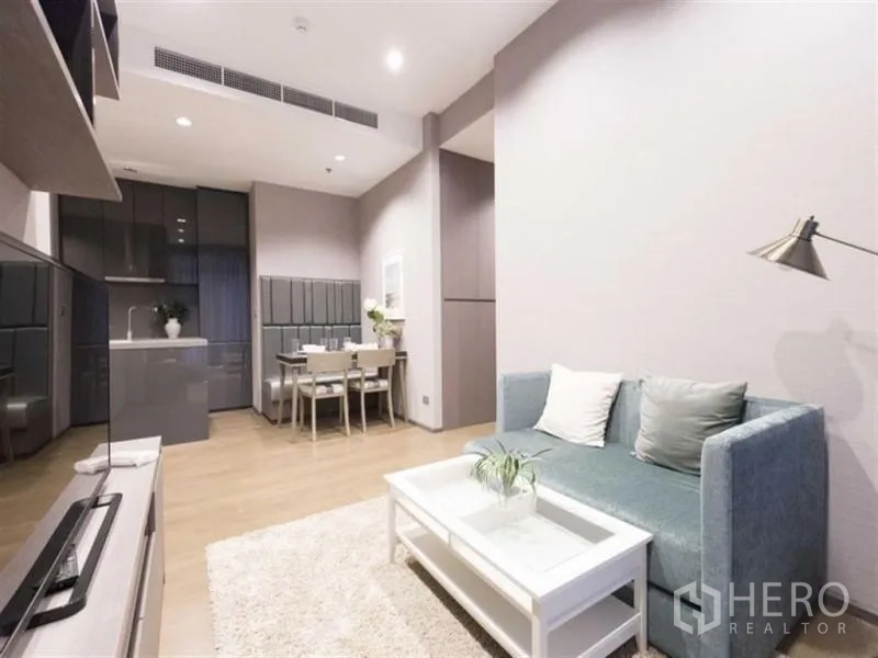 公寓 出租在Bang Rak, Bangkok - 明亮的开放式客厅，青绿色沙发与白色茶几，连通餐区与灰色橱柜厨房，浅木地板。
