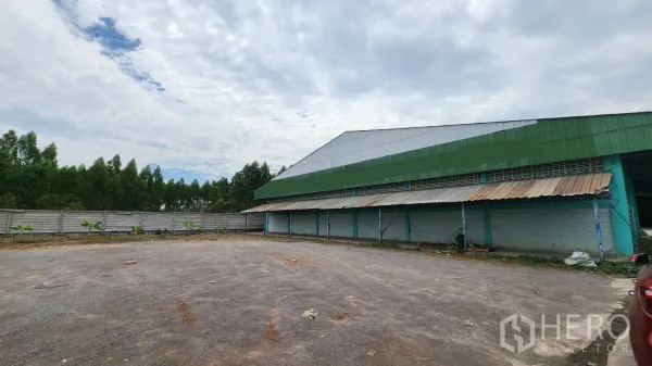 Warehouse for rent in Krathum Baen, Samut Sakhon - Side yard beside the warehouse with a perimeter fence, trees, and a covered loading dock.