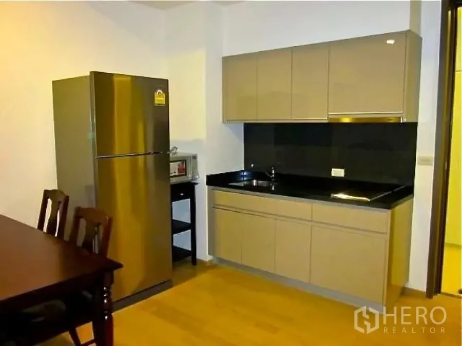 公寓 出租在Watthana, Bangkok - 紧凑厨房，亮面吊柜、黑色台面，配不锈钢冰箱与餐桌椅。