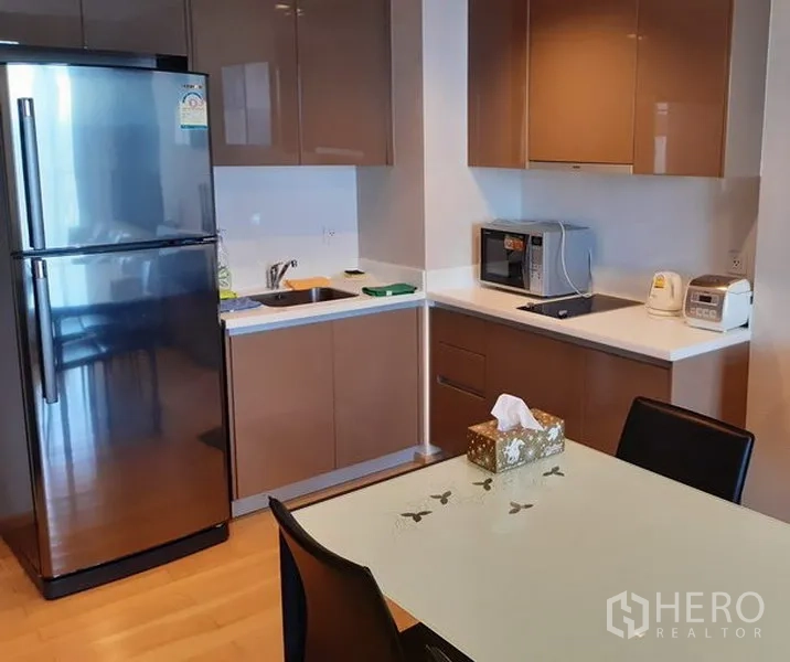 Condominium for rent in Khlong Toei, Bangkok - Modern kitchenette with glossy cabinets, stainless-steel fridge, built-in hob and microwave beside a glass-top dining table.