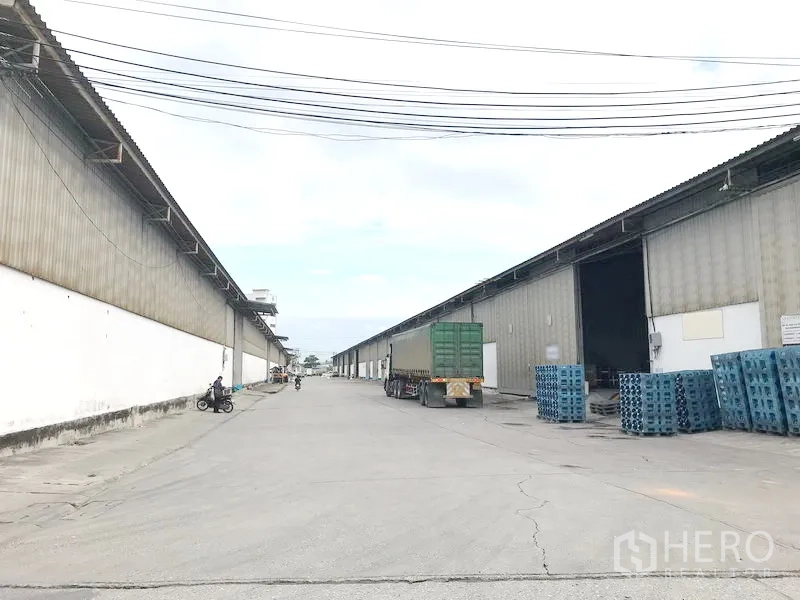 Warehouse for rent in Bang Sao Thong, Samut Prakan - Wide driveway between large corrugated warehouses with loading bays and trucks in Bang Sao Thong.