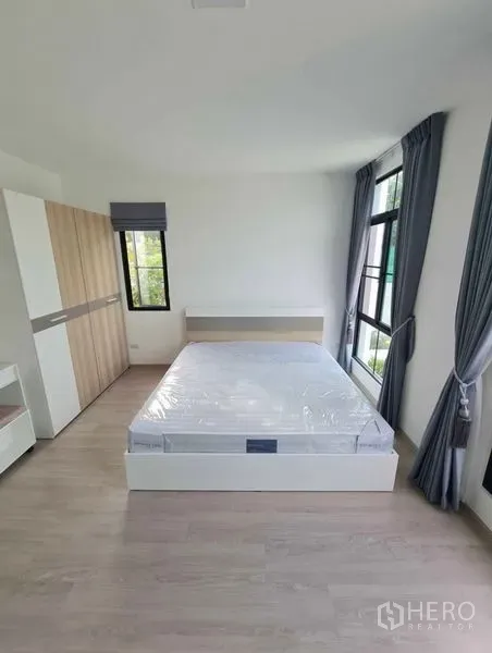 Townhouse for rent in Bang Phli, Samut Prakan - Bright bedroom with double bed, wardrobe and large windows with grey curtains and wood floors.