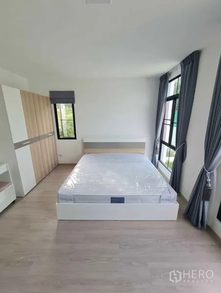 Townhouse for rent in Bang Phli, Samut Prakan - Bright bedroom with double bed, wardrobe and large windows with grey curtains and wood floors.