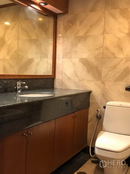 Condominium for rent in Khlong Toei, Bangkok - Bathroom with granite vanity, large mirror and marble-look tiled walls.