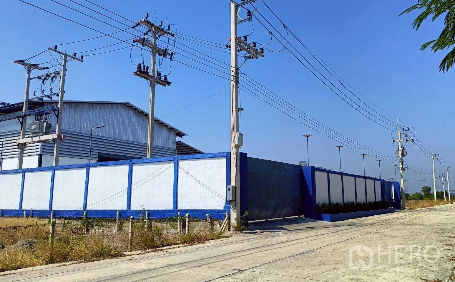 โรงงาน for rent or sale in Mueang Samut Sakhon, Samut Sakhon - รั้วโครงการสีฟ้าขาวสูงพร้อมประตูทางเข้าและเสาไฟฟ้าตามแนวถนน