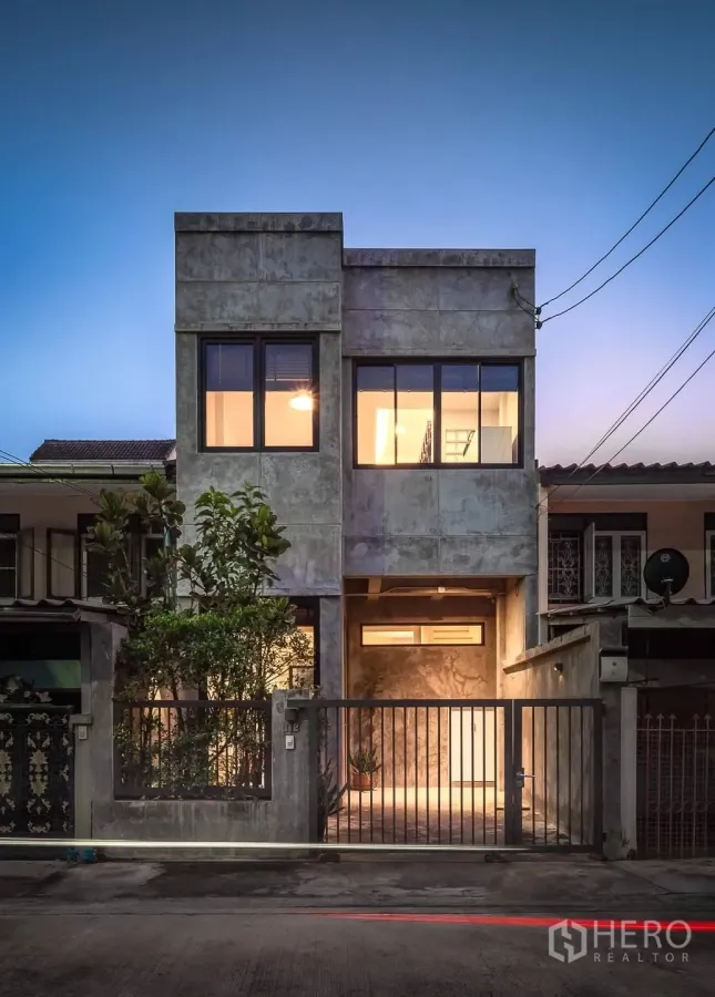 Townhouse for rent in Huai Khwang, Bangkok - Modern concrete-finish townhouse facade at dusk with large windows and gated parking.