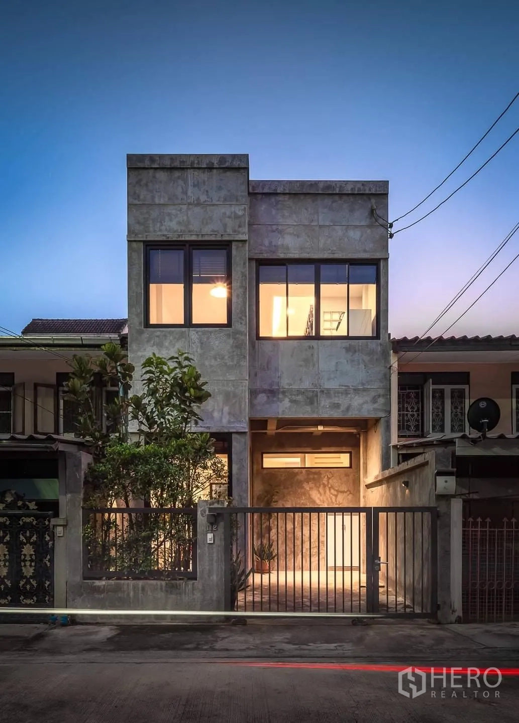 Townhouse for rent in Huai Khwang, Bangkok - Modern concrete-finish townhouse facade at dusk with large windows and gated parking.