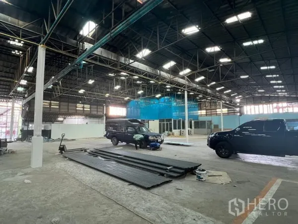 Warehouse for rent in Samut Prakan - Wide warehouse floor under steel trusses with vehicles and stacked metal beams.