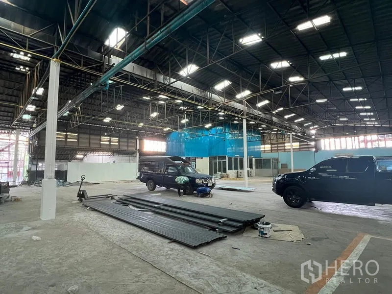 Warehouse for rent in Samut Prakan - Wide warehouse floor under steel trusses with vehicles and stacked metal beams.