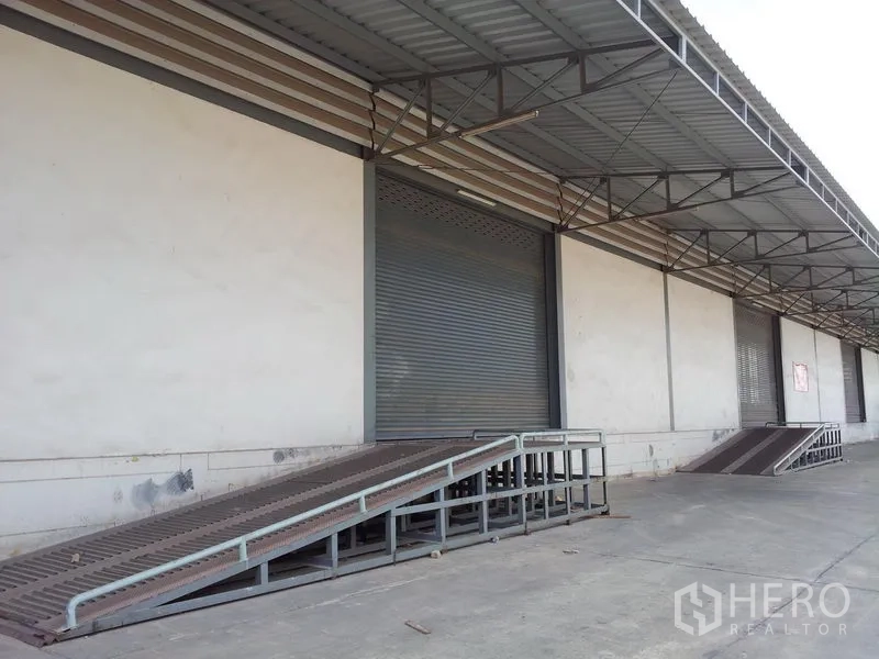 货栈 出租在Mueang Samut Sakhon, Samut Sakhon - 带顶装卸月台、金属坡道与卷闸门的仓库外部，适合卡车进出。