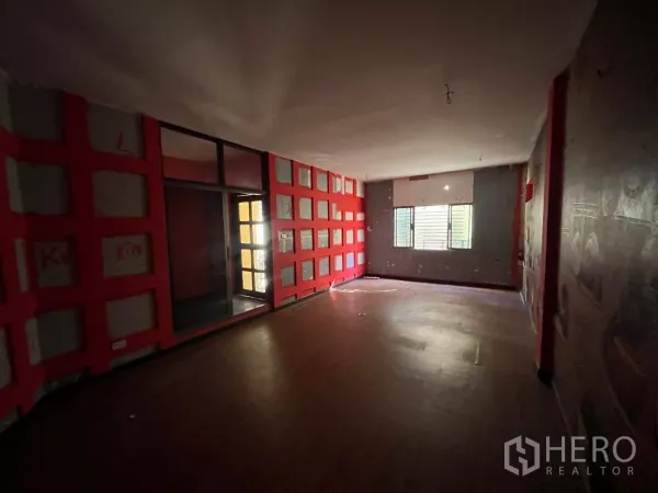 Shophouse for rent in Watthana, Bangkok - Large dim room with a red feature wall, a window with blinds and wood flooring.
