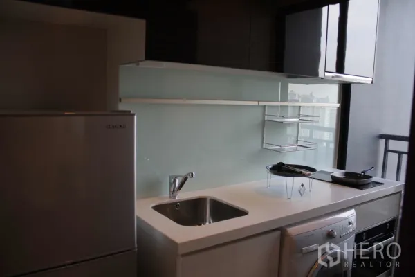 Condominium for rent in Huai Khwang, Bangkok - Compact kitchenette with glossy cabinets, sink, refrigerator, cooktop, oven and washing machine beside a balcony window.