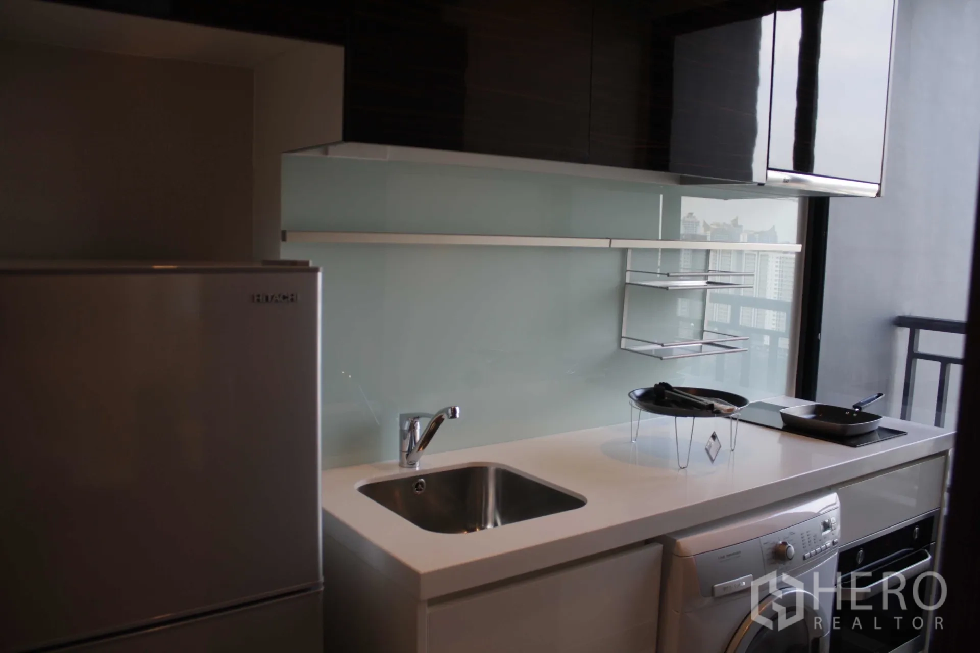 Condominium for rent in Huai Khwang, Bangkok - Compact kitchenette with glossy cabinets, sink, refrigerator, cooktop, oven and washing machine beside a balcony window.