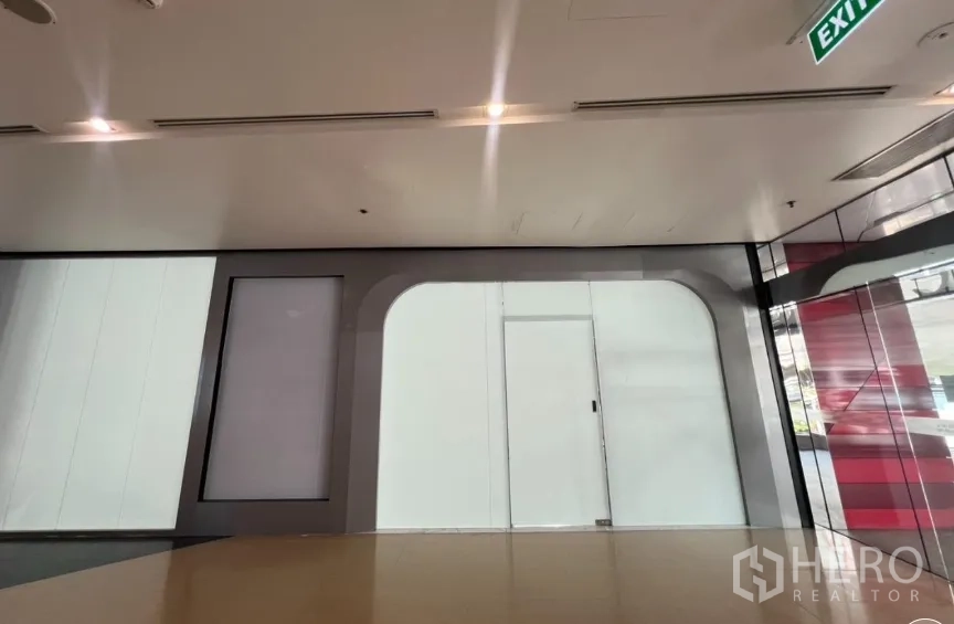 零售空间 for rent in Pathum Wan, Bangkok - 室内走廊旁的玻璃店面，天花灯光与出口标识清晰可见。
