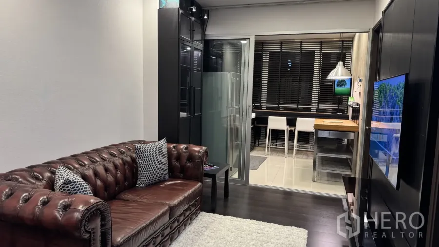Condominium for rent or sale in Khlong Toei, Bangkok - Cozy living room with brown leather Chesterfield sofa, rug, TV wall and a sliding glass door to the kitchen.