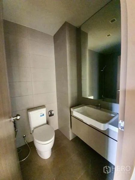 公寓 出售在Ratchathewi, Bangkok - 现代化浴室，带壁挂式洗手台、大镜子和马桶，灰色瓷砖。