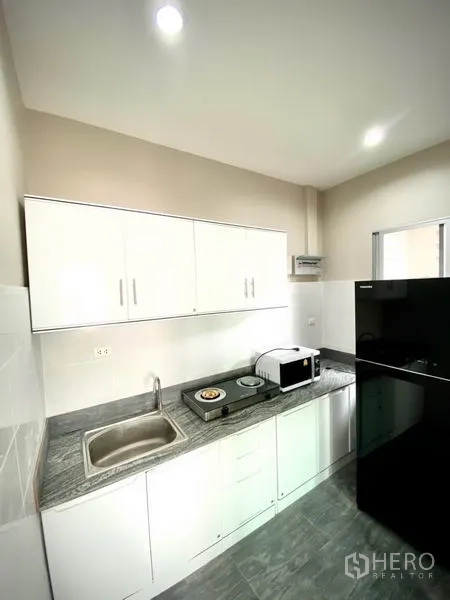 Townhouse for rent in Prawet, Bangkok - Compact kitchen with white cabinetry, stone countertop, sink, cooktop, microwave and large fridge.