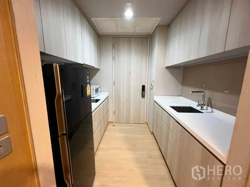 Condominium for rent or sale - Galley kitchen with twin countertops, light wood cabinetry, and black refrigerator.