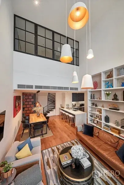Condominium for rent or sale in Sathon, Bangkok - Double-height living room with pendant lights, built-in shelves, leather sofa, dining table, spiral staircase, and open kitchen with breakfast bar.