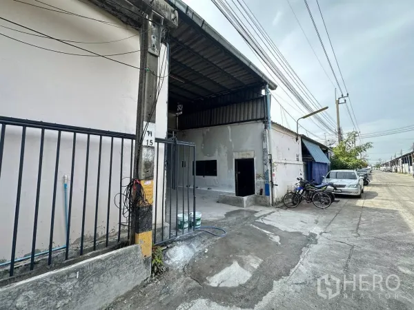 工厂 出租在Samut Prakan - 工厂外立面与滑动大门，临工业小路，可路边停车并有电线杆。