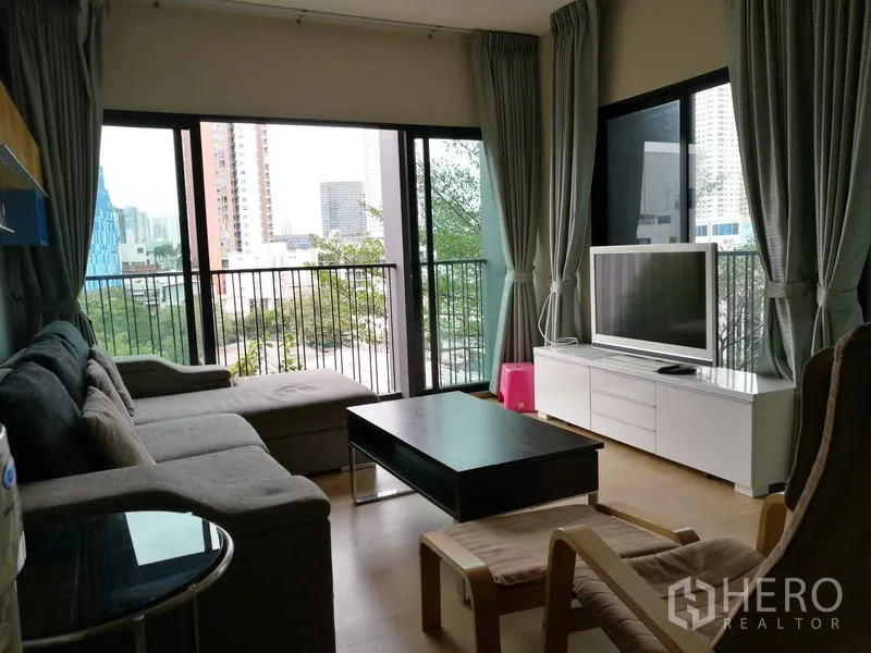 คอนโด ให้เช่าในWatthana, Bangkok - ห้องนั่งเล่นโซฟาเข้ามุม โต๊ะกลาง ทีวีบนตู้สีขาว หน้าต่างรอบด้านเปิดสู่ระเบียง วิวเมือง