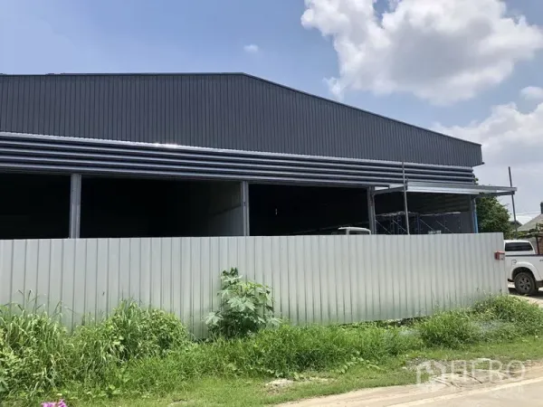 คลังสินค้า ให้เช่าในBang Khen, Nonthaburi - มุมมองด้านข้างของโกดังโลหะขนาดใหญ่ หลังรั้วชั่วคราวและริมสนามหญ้า