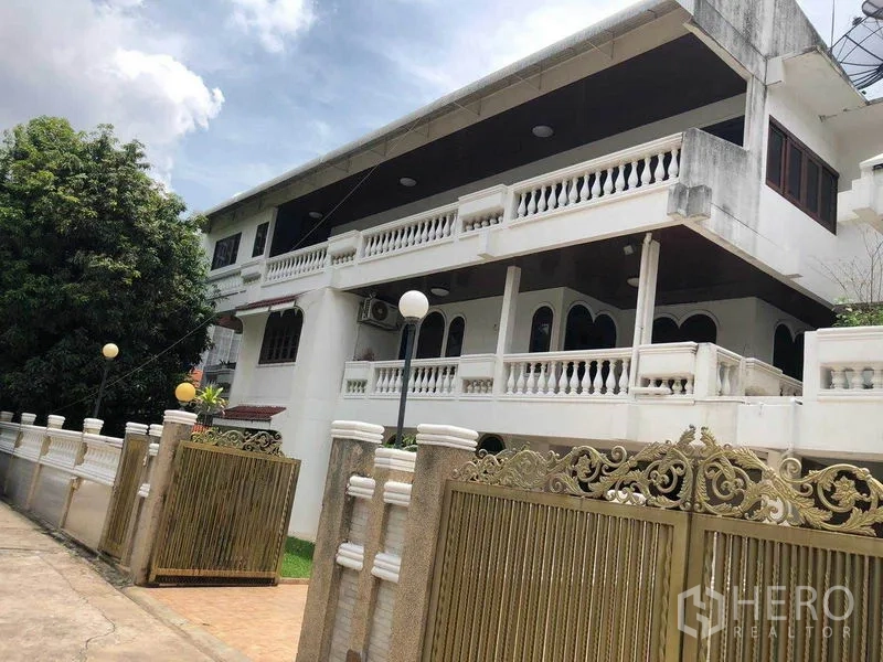 Detached House for rent in Watthana, Bangkok - Two‑storey detached house with white balustrades and ornate golden gate under a cloudy sky.