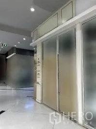 零售空间 出租在Khlong Toei, Bangkok - 走廊视角的零售店面，磨砂玻璃立面、金属把手、嵌入式顶灯与白色地面。