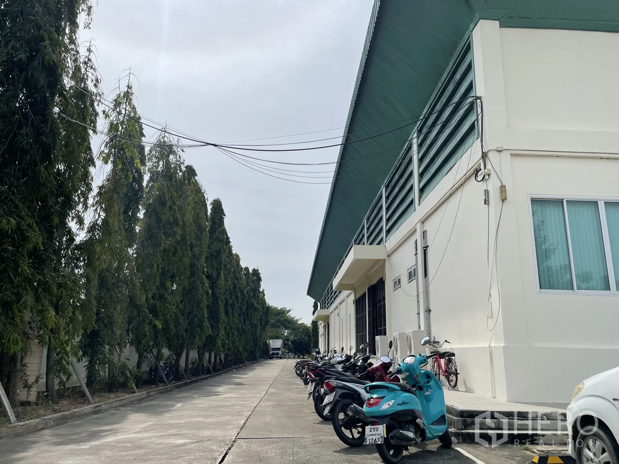 Warehouse for sale in Lat Lum Kaeo, Pathum Thani - Tree‑lined side lane with motorcycle parking and service doors along the warehouse.