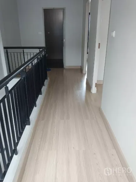 Townhouse for rent in Mueang Samut Prakan, Samut Prakan - Bright upstairs hallway with light wood flooring, black railing, and a bathroom door.