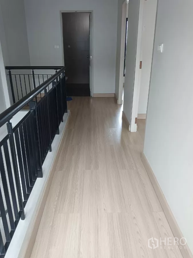 Townhouse for rent in Mueang Samut Prakan, Samut Prakan - Bright upstairs hallway with light wood flooring, black railing, and a bathroom door.