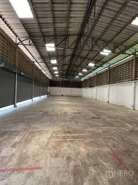 Warehouse for rent in Bang Na, Bangkok - Open-plan warehouse interior featuring a high steel-truss roof, skylights, and concrete flooring with roller shutters along one side.